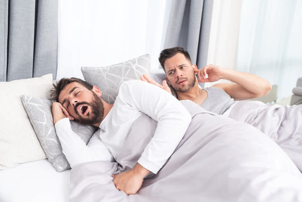 Frustrated man blocking his ears while his partner snores in bed beside him