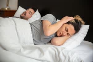 Is Snoring Bad? A woman looks frustrated in bed as her partner snores loudly, highlighting sleep disturbance issues.